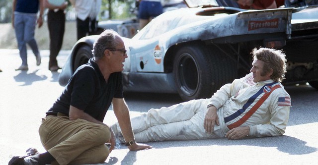 A man with glasses speaking to a racing pilot laying on the floor