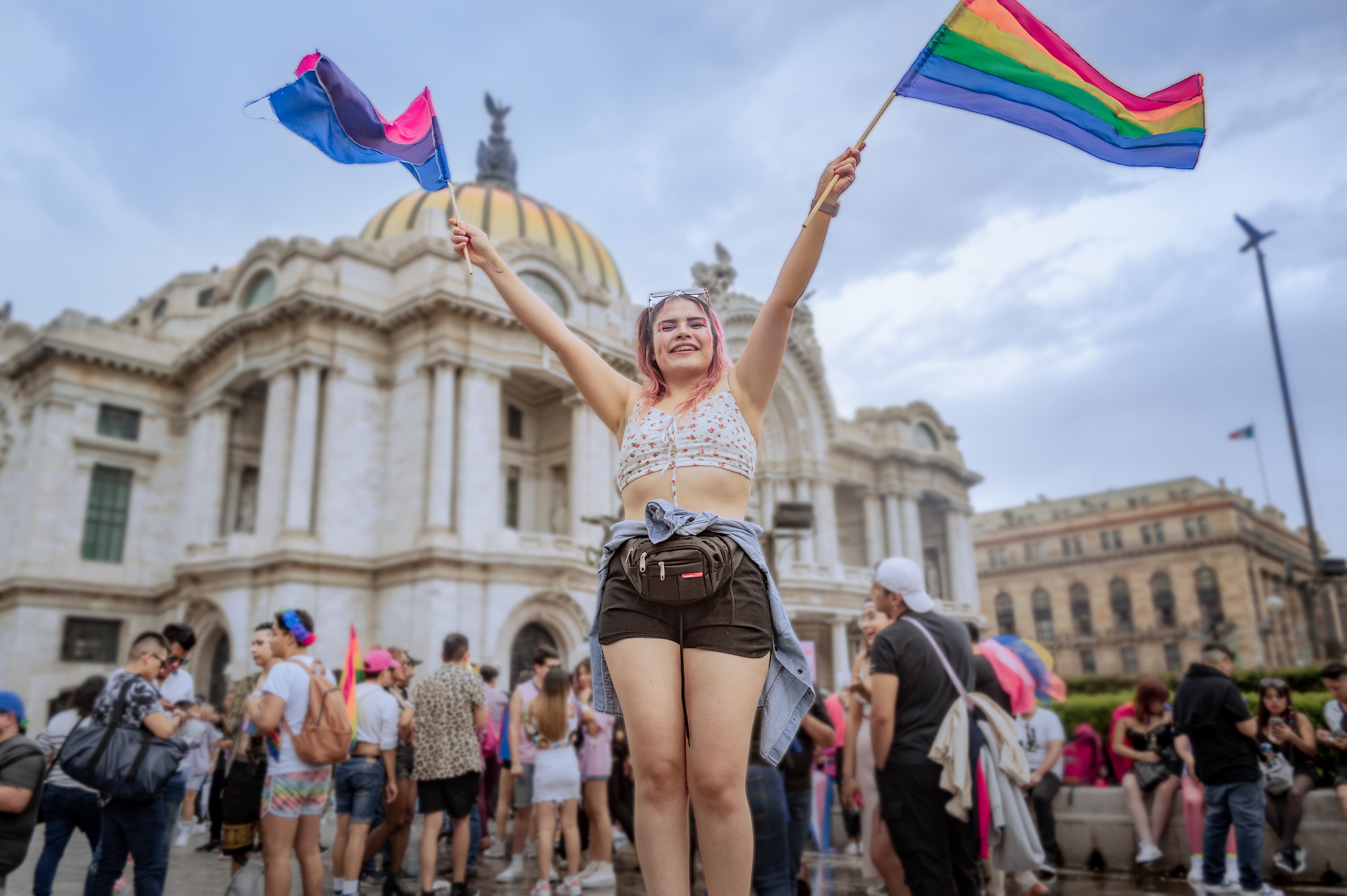 A pink haired woman holding a LGBT flag up in her left hand and a bisexual flag in her right hand