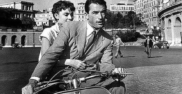 A man dressed in a suit rides a motorcycle, he has a woman behind him