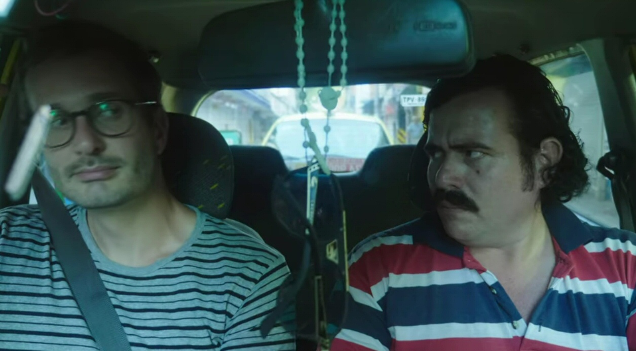 Inside a car, a man with dark hair and glasses, and a man with short hair and a mustache.
